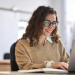 Young happy woman employee or student sitting at desk using laptop.